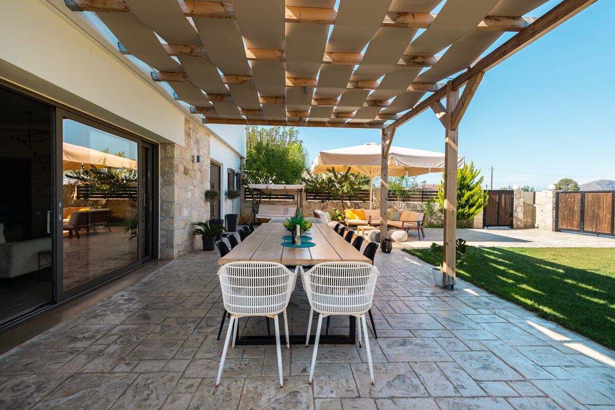 An outdoor dining area is presented, featuring a long table surrounded by multiple white chairs. A wooden pergola provides shade overhead, with soft fabric panels filtering sunlight. The space opens to a well-manicured lawn and showcases a serene ambiance for gatherings.