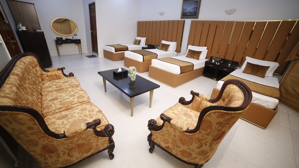 A spacious room is arranged with multiple beds, showcasing a blend of modern and traditional decor. Luxurious seating is provided by elegantly patterned chairs and a central coffee table, creating a comfortable social area for guests. Natural light fills the space, enhancing its warm tones.