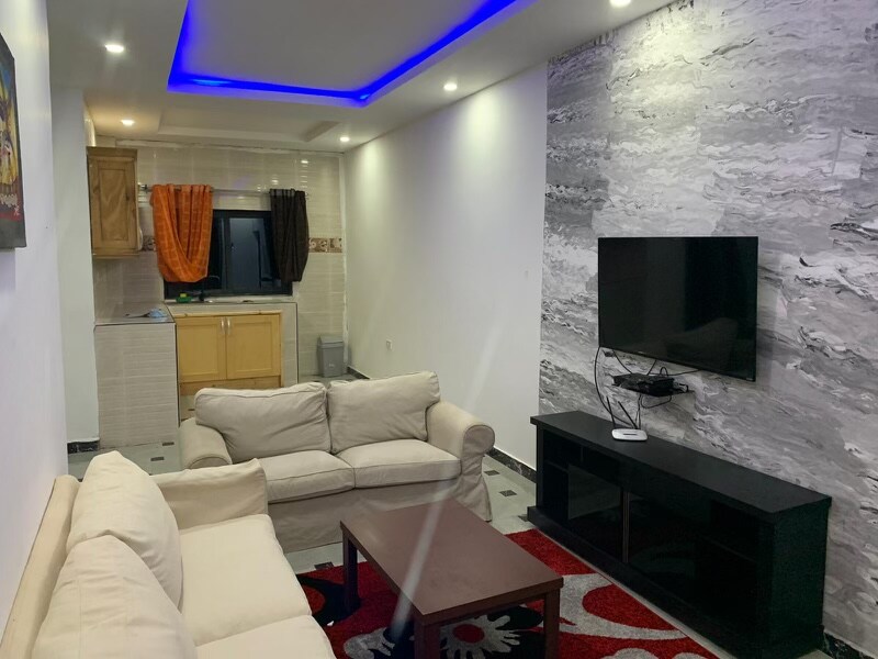 A cozy living area features a cream-colored sofa set arranged around a dark wood coffee table. A modern TV stands on a black console against a textured wall. Soft lighting creates a relaxed ambiance, while a glimpse of a kitchen area is visible in the background.