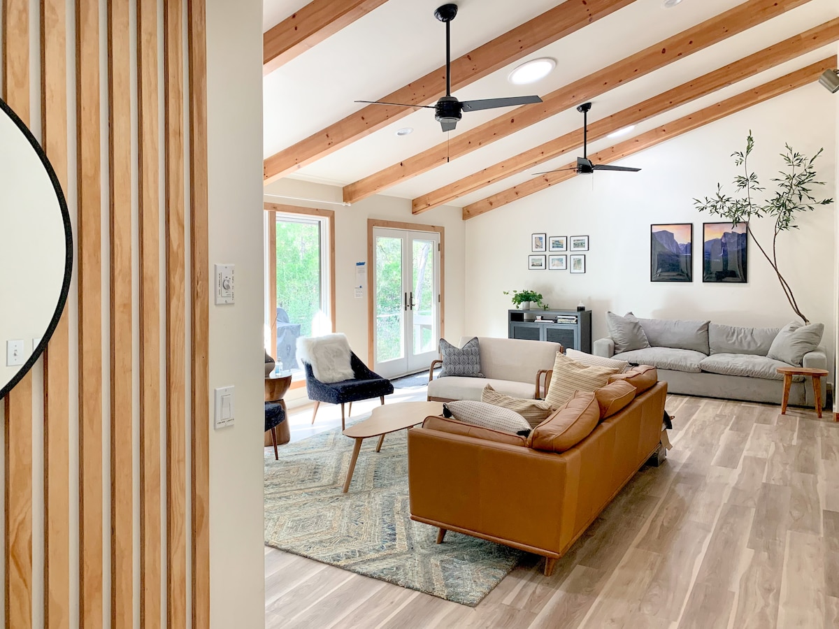 The living room features vaulted ceilings with exposed wooden beams. Abundant natural light floods through large windows, illuminating the spacious seating area, which includes a leather sofa, armchairs, and a patterned rug. A corner cabinet and decorative wall art add interest to the cozy space.