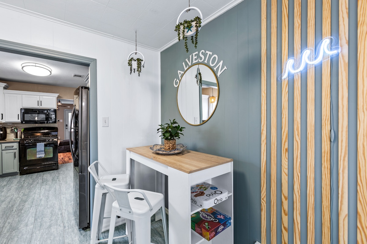 A modern dining area features a light wood countertop with two white stools. A round mirror with 'GALVESTON' written above is mounted on a wall, alongside decorative wood panels and playful neon wave signage. Potted plants add a refreshing touch.