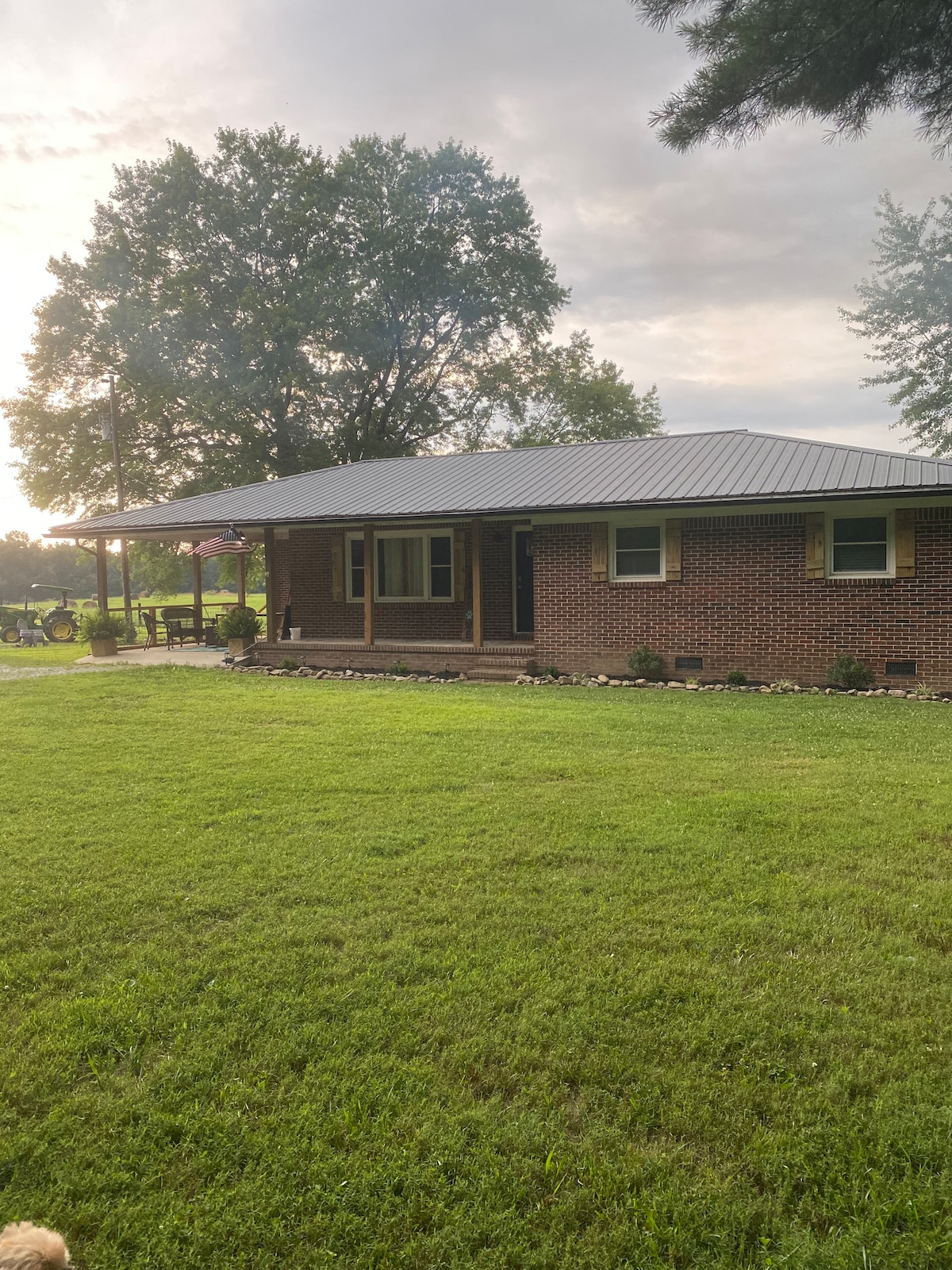 A one-story farmhouse is shown with a brick exterior and a metal roof. The front porch is framed by decorative shutters, and green grass surrounds the home. Large trees provide shade, and a flagpole stands nearby, adding to the welcoming atmosphere.