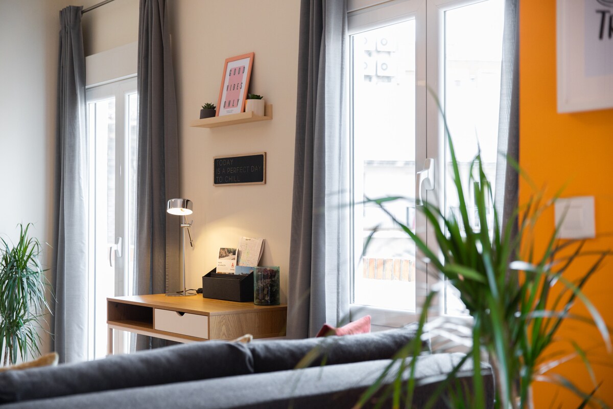 A well-lit corner of the living room features a modern desk with a lamp and decorative items. Two tall windows are dressed with gray curtains, allowing natural light to fill the space. Green plants add a touch of nature to the setting.