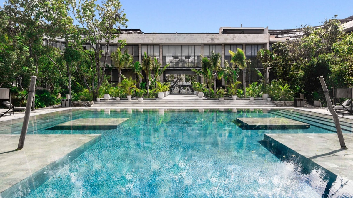 A large swimming pool is surrounded by lush greenery and palm trees. The water reflects sunlight, creating a serene atmosphere. Lounge chairs are positioned around the pool, providing spaces for relaxation. The background features modern architecture, enhancing the inviting outdoor setting.
