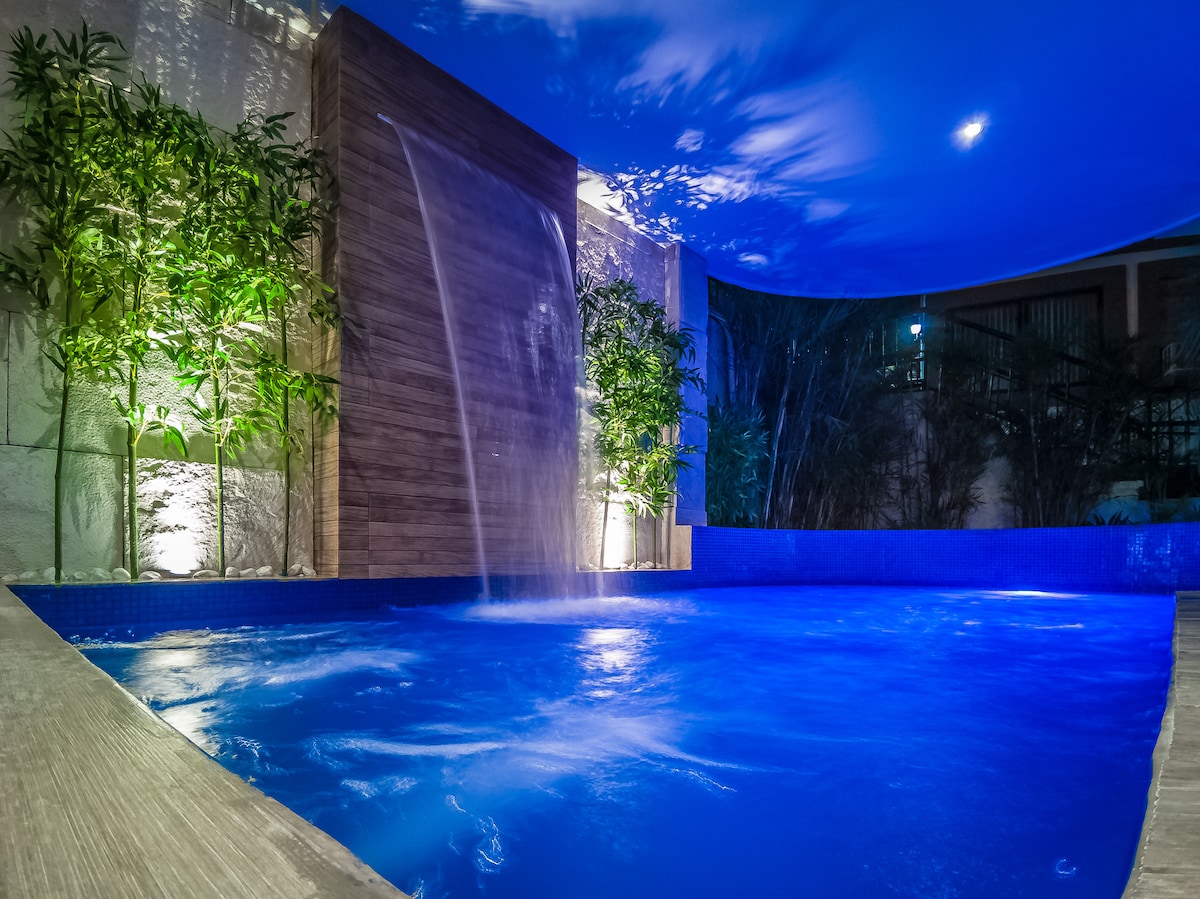 The private pool area features a soothing waterfall that cascades into the bright blue water. The space is surrounded by lush greenery, contributing to a serene ambiance. Soft lighting enhances the calming atmosphere, making it an inviting spot for relaxation.