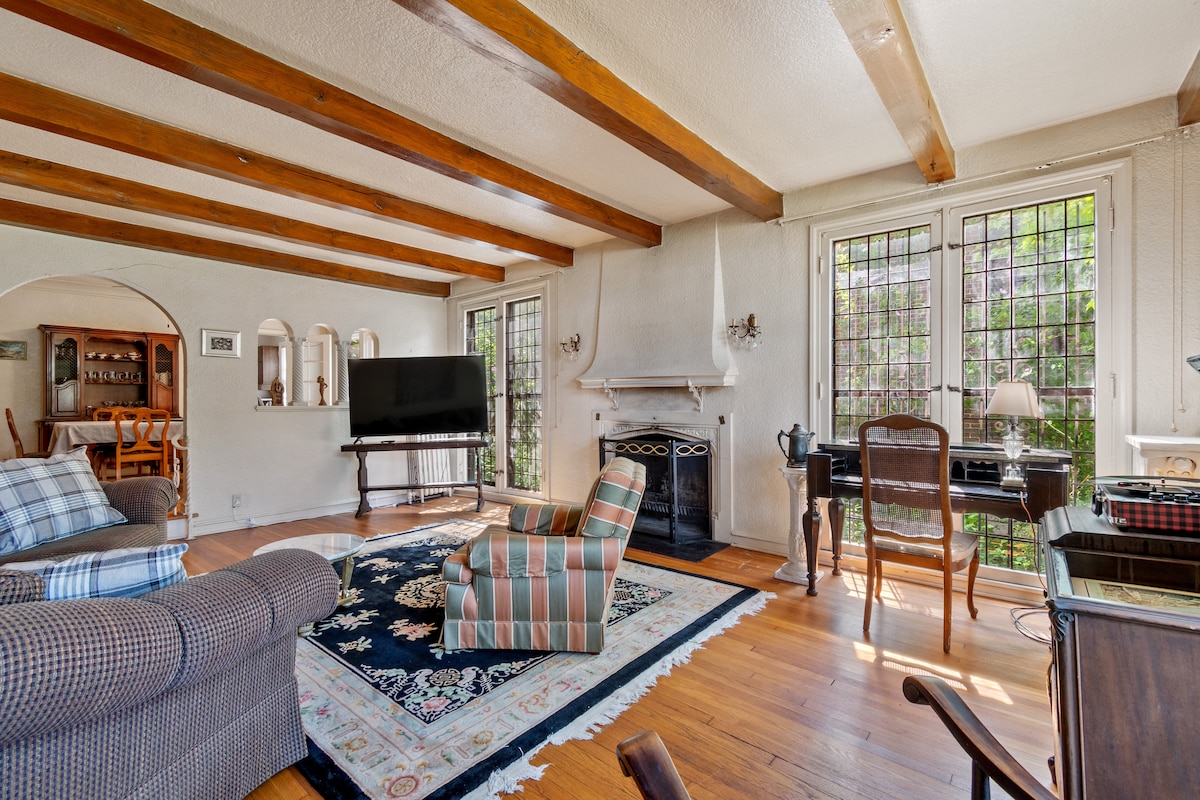 An inviting living area features exposed wooden beams along the ceiling, with a cozy seating arrangement that includes a striped chair and a plush sofa. A decorative rug covers the hardwood floor, while large windows provide natural light, framed by leafy greenery outside.