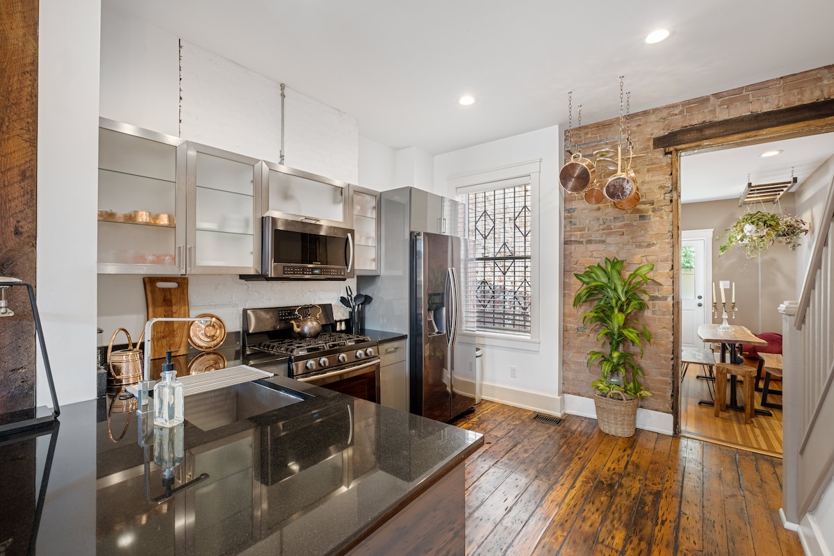 The kitchen features sleek stainless steel appliances and granite countertops, complemented by wooden flooring. A large window allows natural light, while a decorative plant adds a touch of greenery. An open doorway leads to a dining area, enhancing the flow of space.