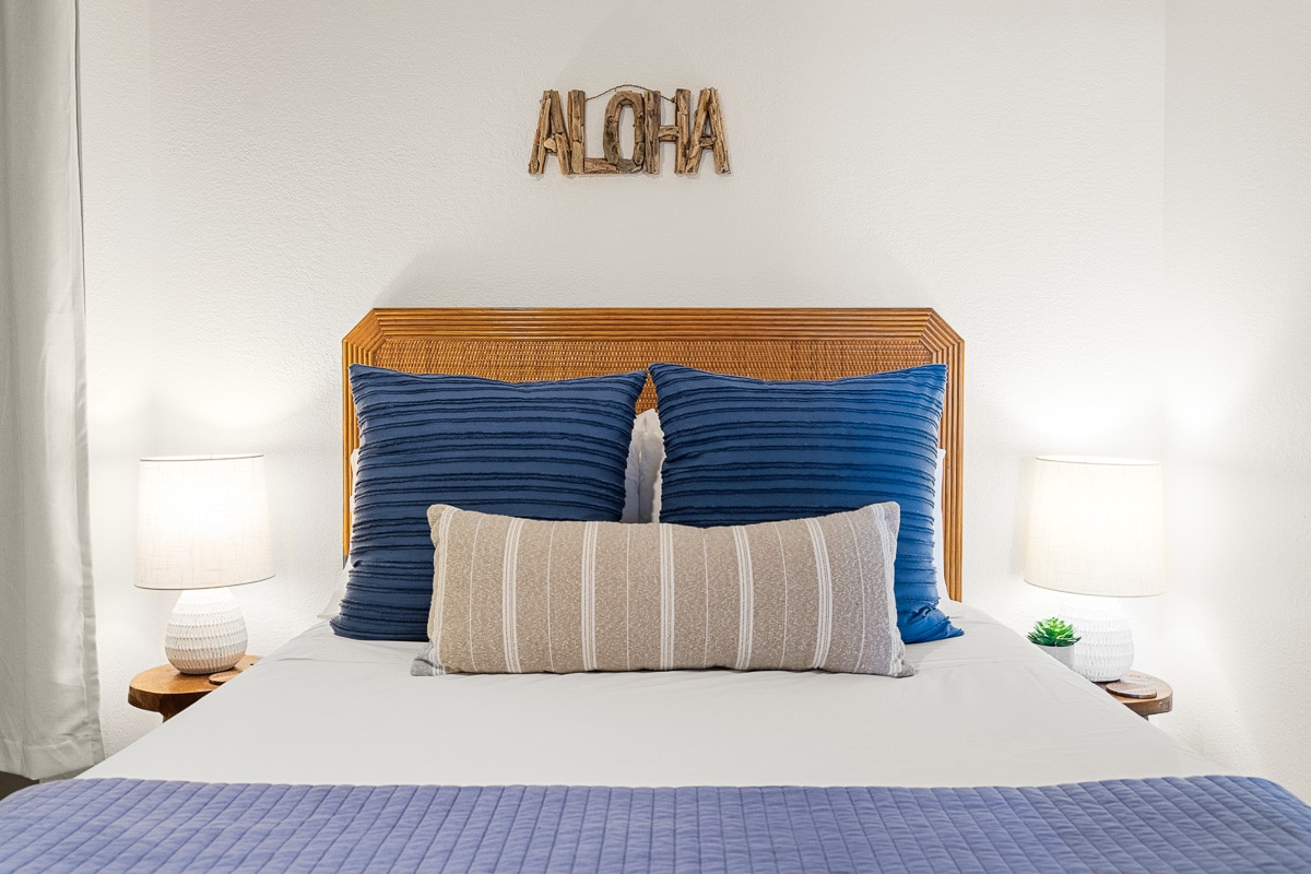 A comfortable bedroom features a spacious bed with navy blue and neutral pillows. A decorative 'Aloha' sign is displayed above the headboard. Soft lighting from two bedside lamps creates a calming atmosphere.
