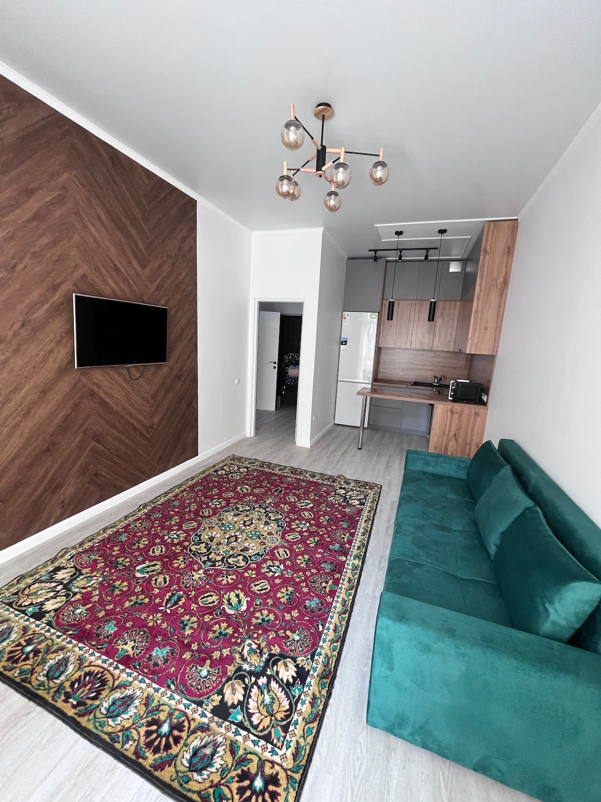 A spacious living area features a decorative area rug with intricate patterns on a light floor. A teal sofa is positioned against a textured feature wall, and a modern light fixture hangs from the ceiling. An open kitchen area is visible in the background.