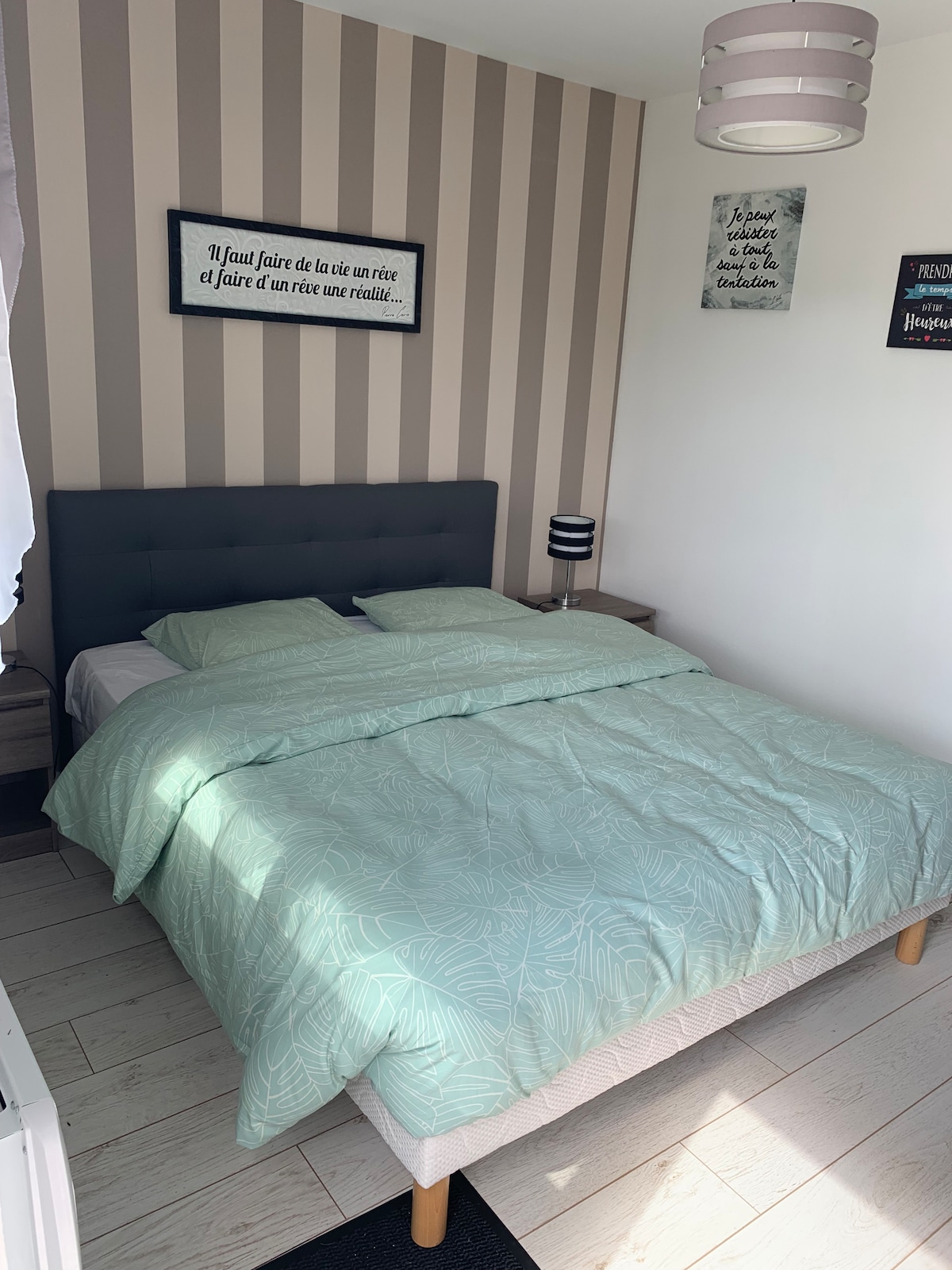 A cozy sleeping area is presented with soft, mint green bedding arranged neatly on a spacious bed. The walls are adorned with light and dark striped patterns, complemented by decorative wall art. A bedside table with a lamp sits next to the bed, providing a functional touch.