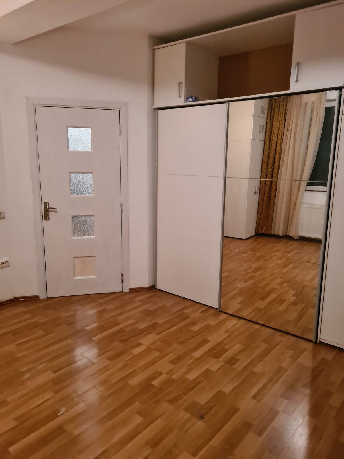 A bright room is presented, featuring a smooth wooden floor and a mirrored wardrobe that reflects the space. A white door with glass panels is visible, along with floor-to-ceiling curtains that frame the window, contributing to a sense of openness.