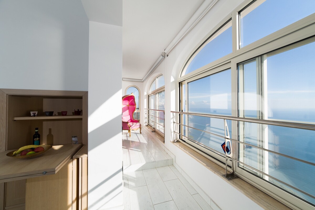A light-filled space features large arched windows showcasing panoramic sea views. A simple wooden cabinet is positioned to the left, with bottles and decorative items arranged neatly. A vibrant pink chair is visible in the corner, inviting relaxation while taking in the coastal scenery.