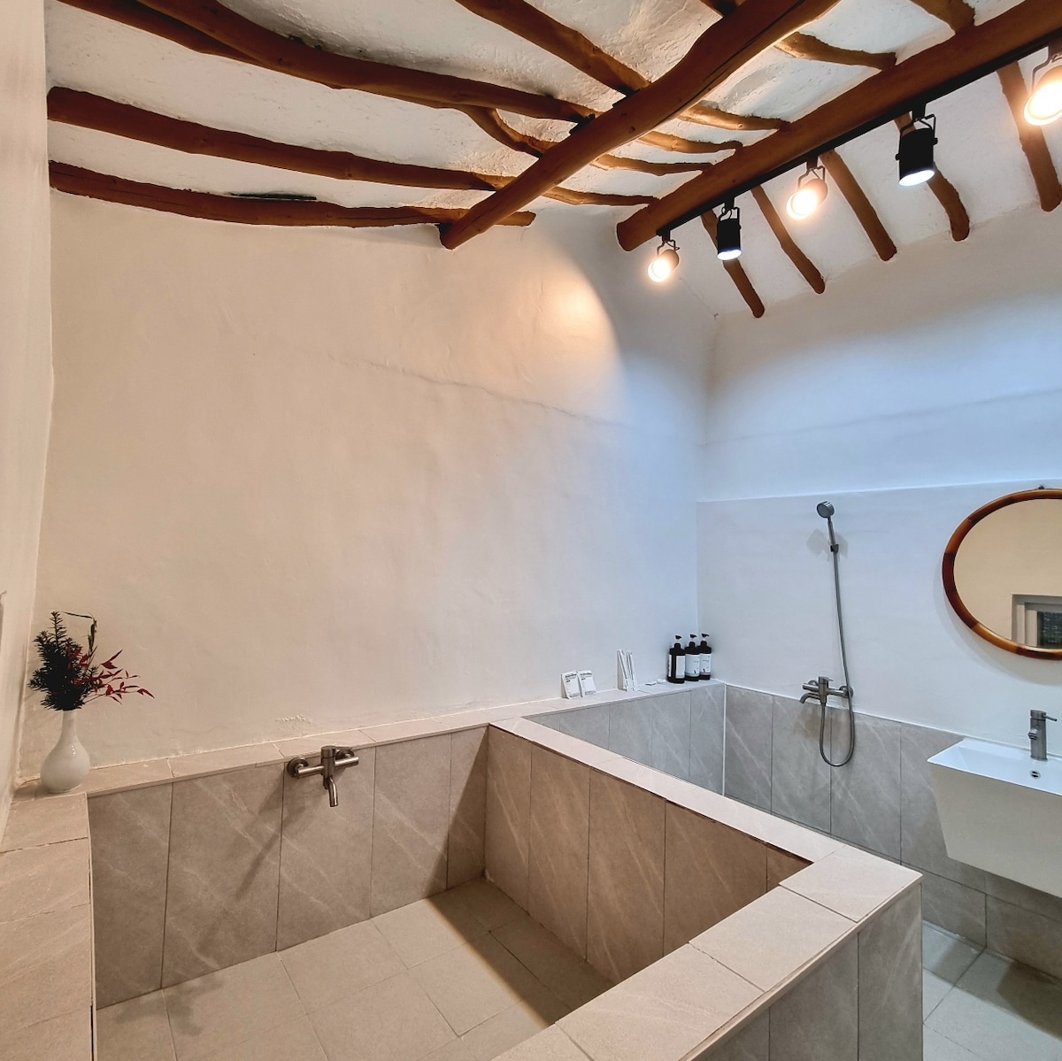 A spacious bathroom features a unique sunken tub with a sleek faucet and an overhead shower. Warm lighting highlights the white walls and rustic wooden beams above. A circular mirror is positioned above a modern sink, with neatly arranged bottles and a small vase adding a touch of decor.