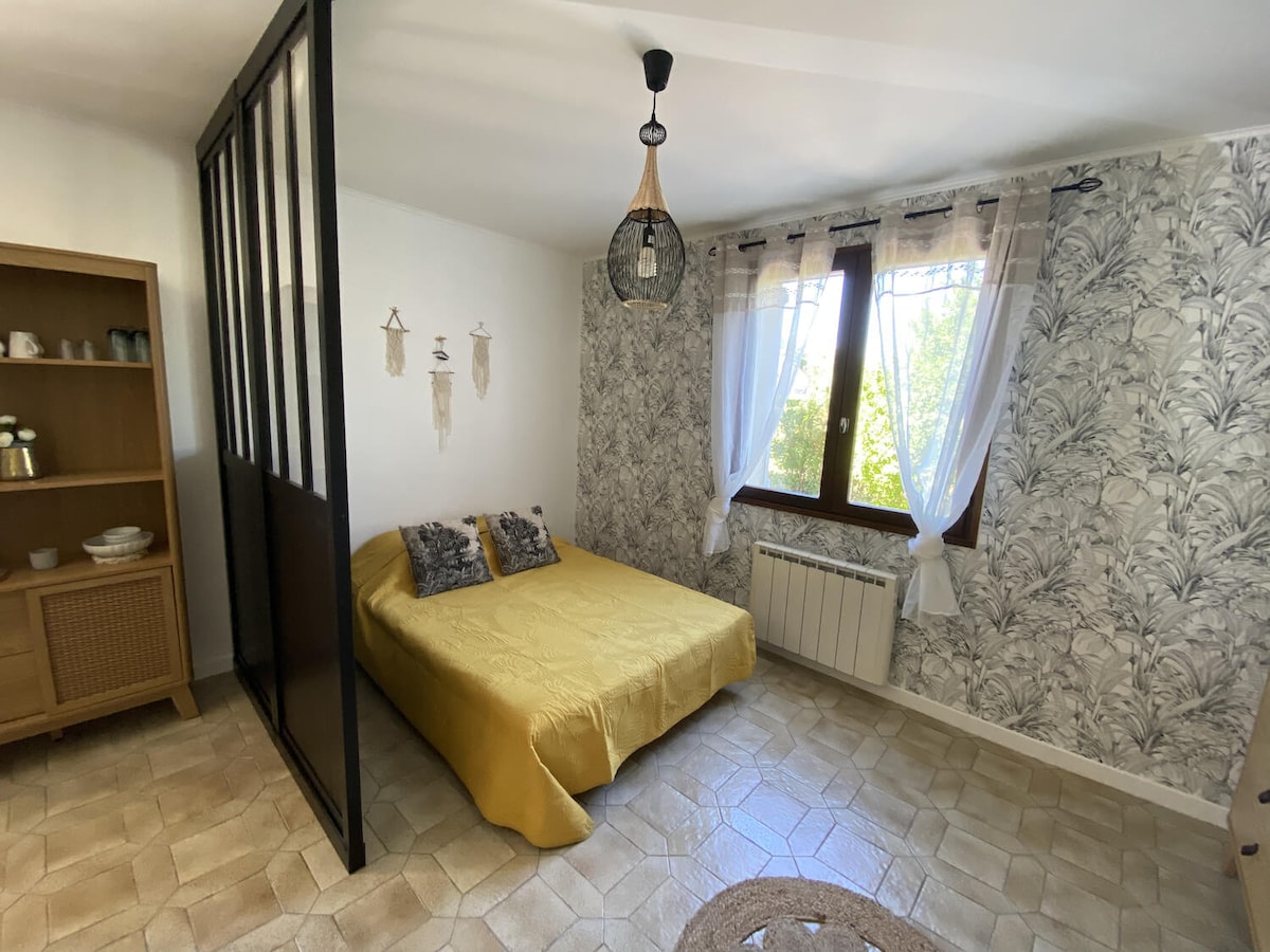 A cozy sleeping area features a spacious bed dressed in a yellow coverlet. Natural light enters through two windows adorned with light curtains, illuminating the patterned wallpaper. A decorative shelf holds tableware in the corner, and a light fixture adds subtle charm.