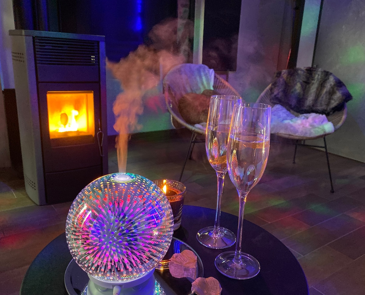 A cozy indoor space is illuminated by soft, colorful lighting. A decorative diffuser sits on a table, producing a gentle mist. Two glasses are ready for a toast, and a warm fireplace adds a comforting ambiance. Two stylish chairs are positioned nearby.