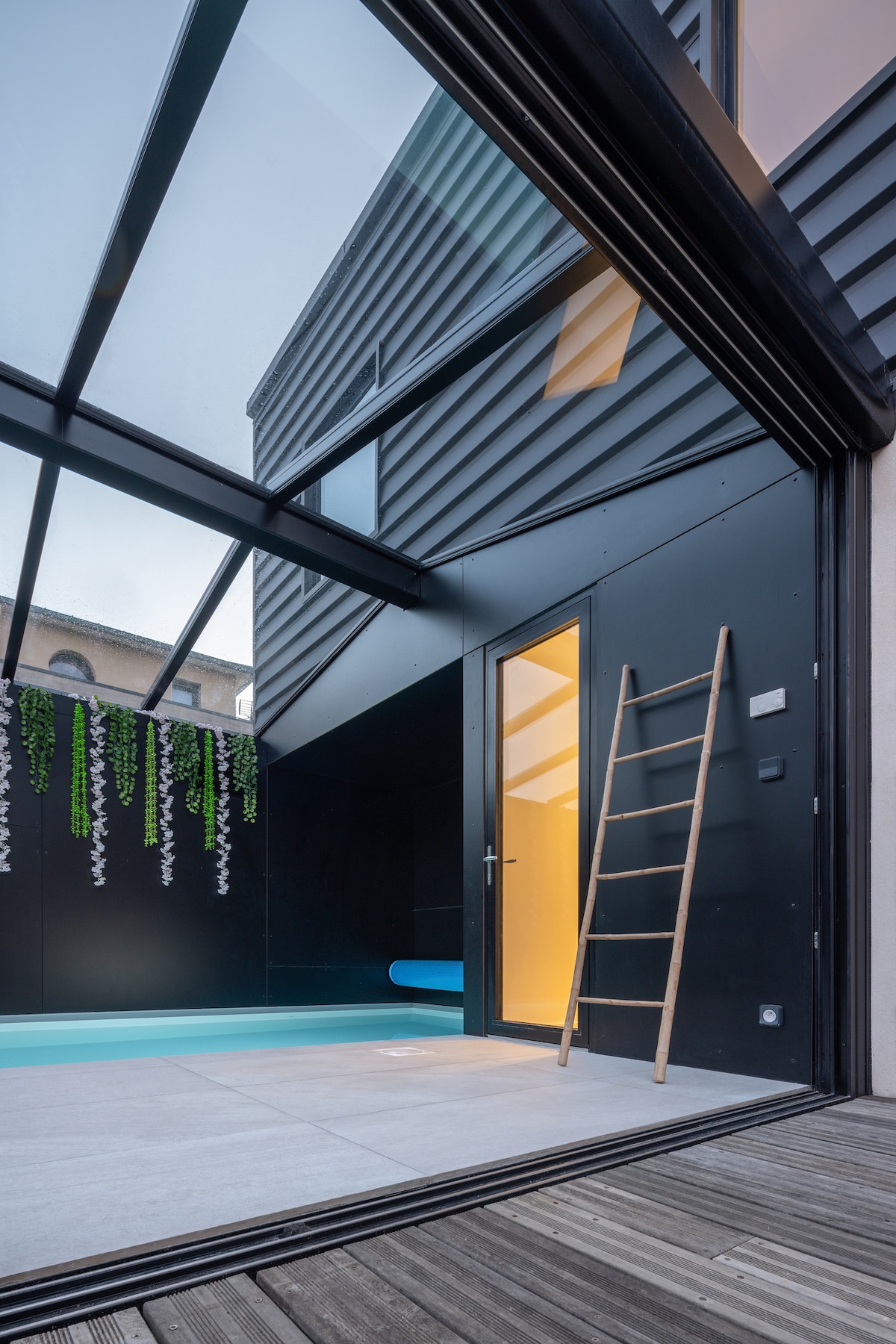 A modern indoor pool area features a large glass roof, allowing natural light to illuminate the space. The pool is surrounded by smooth tiles, with a wooden ladder positioned against the wall. Greenery cascades from the walls, enhancing the serene ambiance.