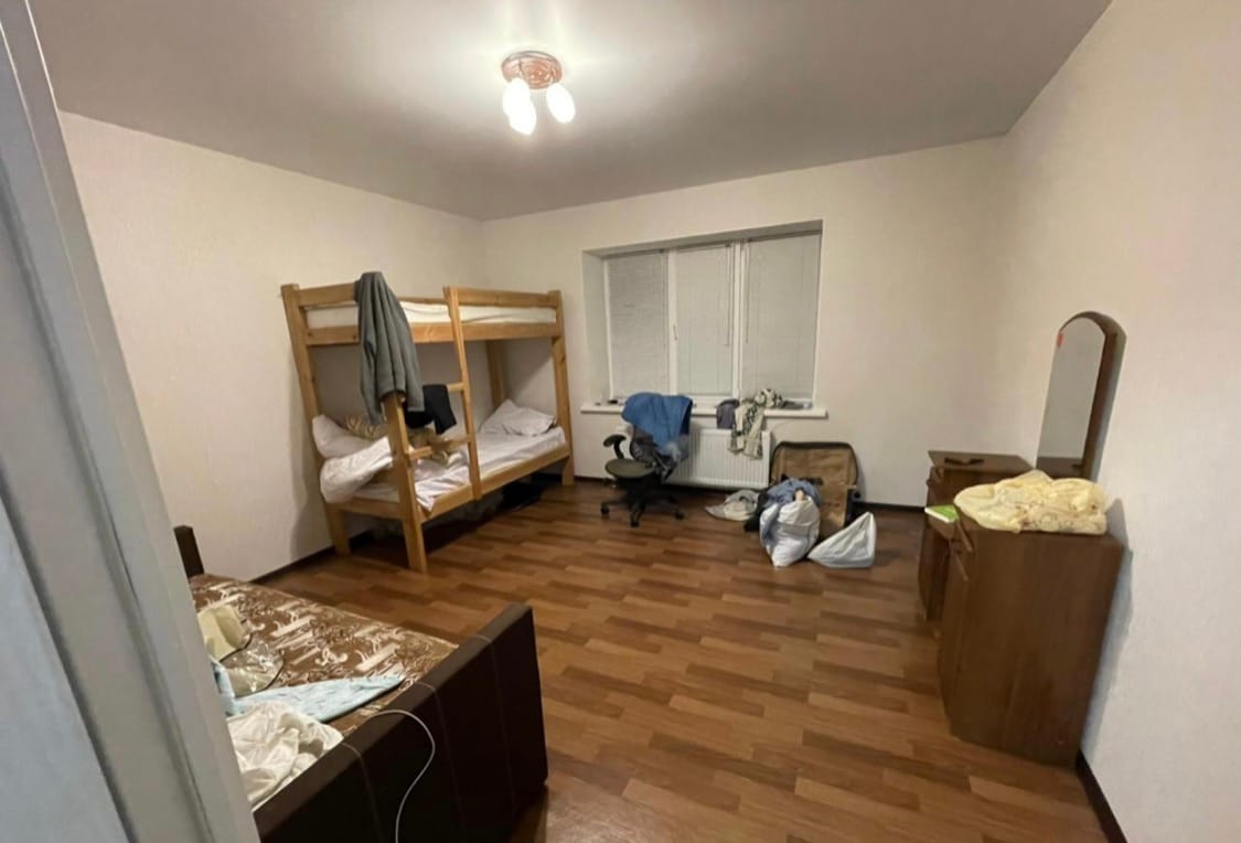 The room features a wooden bunk bed with two twin-sized mattresses. A desk chair is positioned next to a window, which allows natural light to enter. A dresser is present along one wall, and various items are scattered throughout the space.