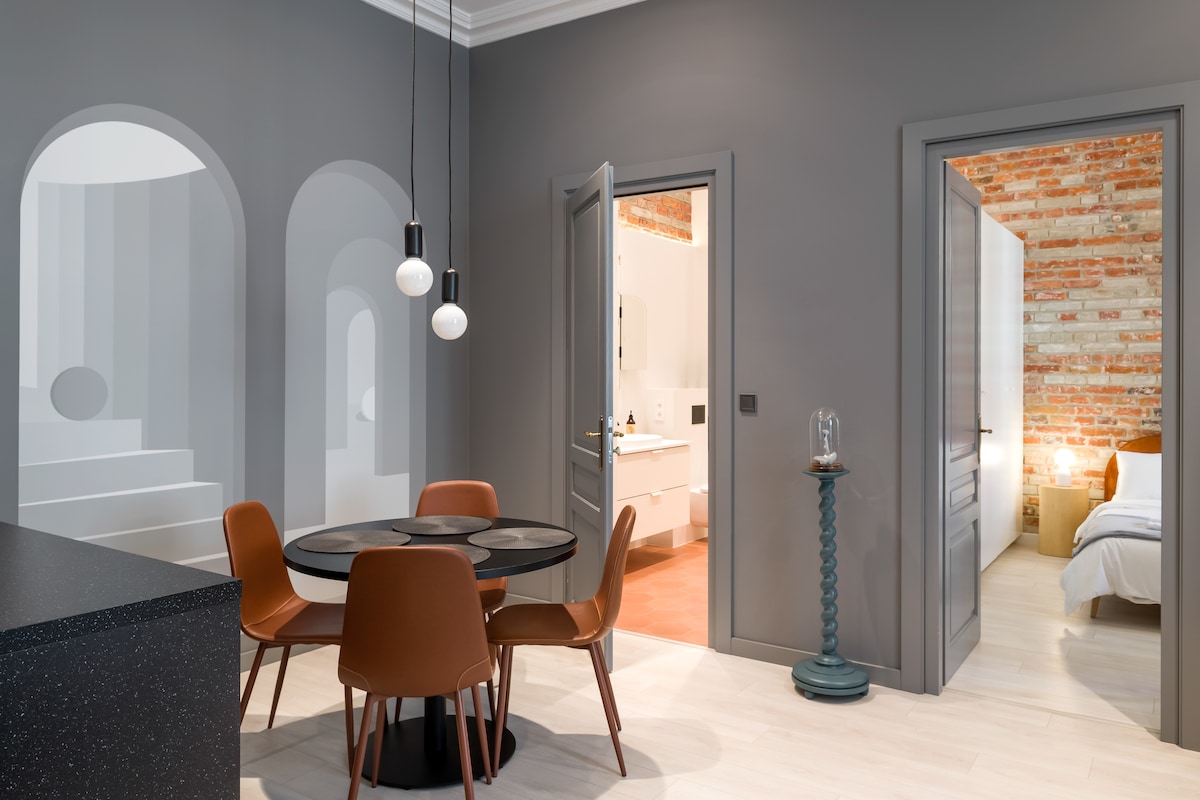 A sleek dining area features a round table surrounded by four mid-century modern chairs. Two minimalist pendant lights hang above, illuminating the space. In the background, the doorway leads to a stylishly designed bedroom, while a textured brick wall adds character to the ambiance.