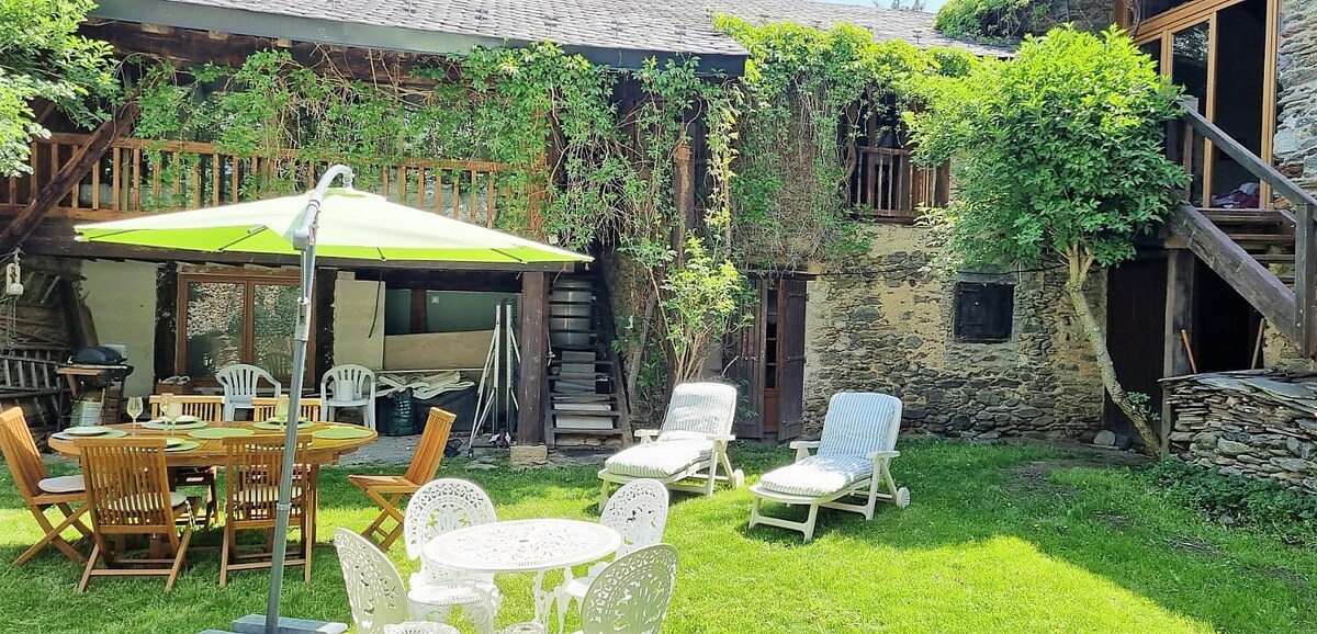 The outdoor area features a sunny garden with a mix of wooden and white furniture, including lounge chairs and a dining table. A large green umbrella provides shade over the seating area. Stone walls and climbing greenery frame the space, creating a tranquil retreat.