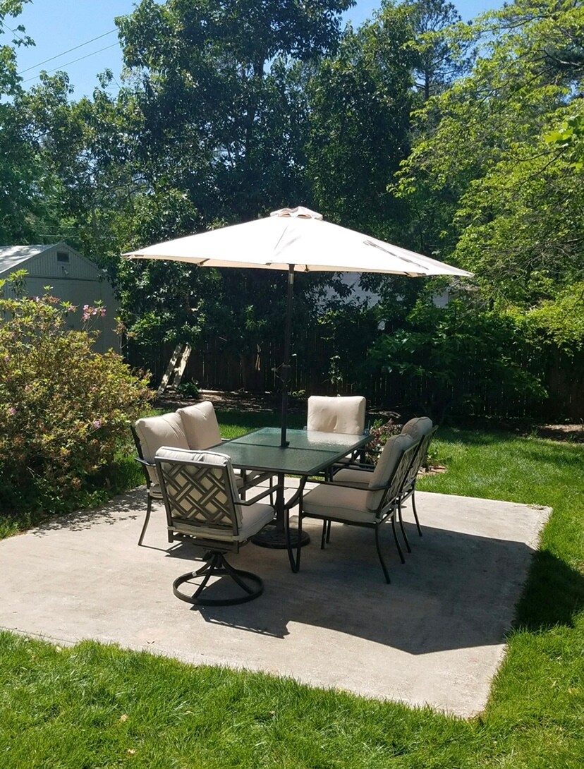 A shaded outdoor dining area features a glass table surrounded by six chairs. An umbrella provides protection from the sun, while lush greenery and blooming plants add a natural touch to the space. A concrete patio serves as the foundation for this inviting setting.