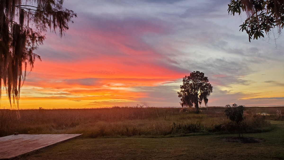 A vibrant sunset casts colorful hues across the sky, reflecting on the tranquil waters of Lake Istokpoga. Tall grasses and a solitary tree frame the scene, while a wooden deck offers a perspective on this serene natural setting.