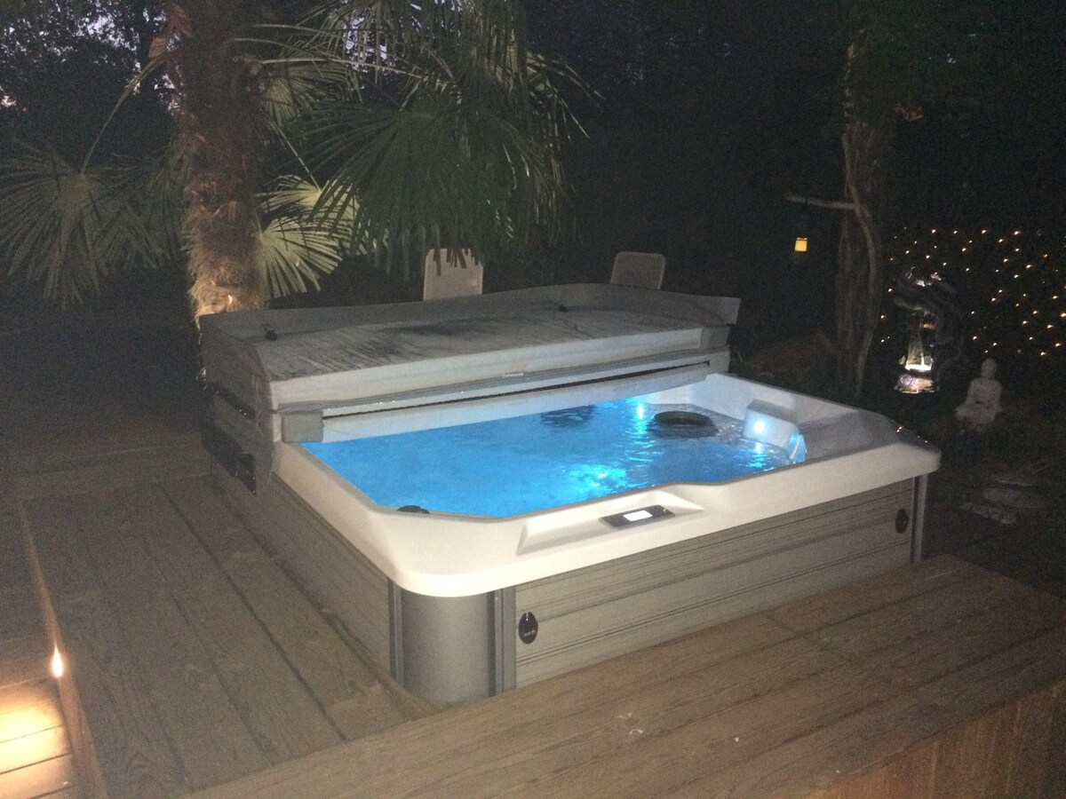 A private outdoor spa is situated on a wooden deck, surrounded by lush greenery. Soft lighting illuminates the area, highlighting the spa's blue water and providing a relaxing ambiance. The spa cover is partially closed, and comfortable seating is visible nearby.