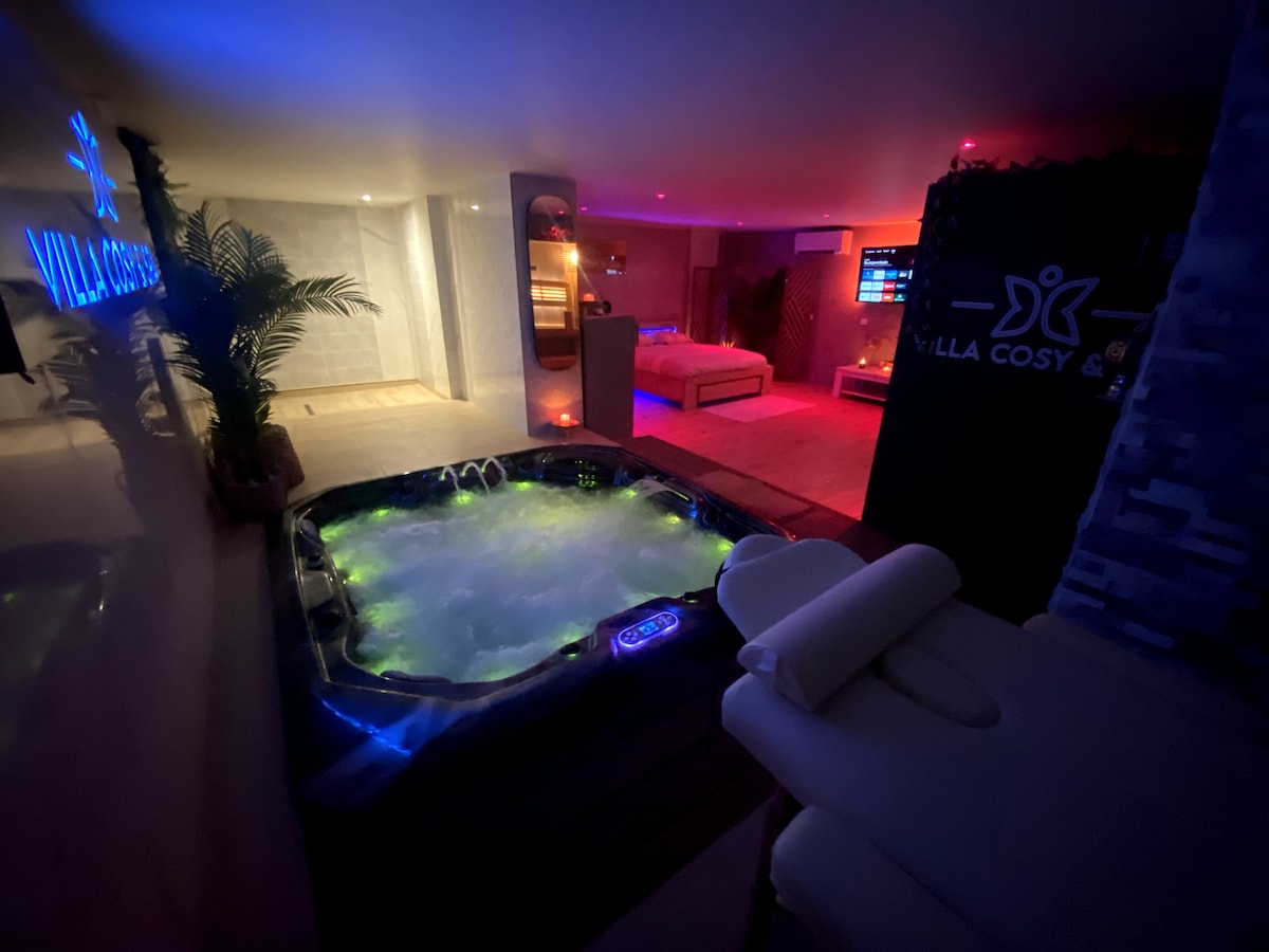 An indoor spa area is illuminated with ambient lighting in various colors. A jacuzzi is filled with bubbling water, surrounded by soft lighting. A massage table is positioned nearby, with towels neatly arranged on top, creating a serene and relaxing environment.