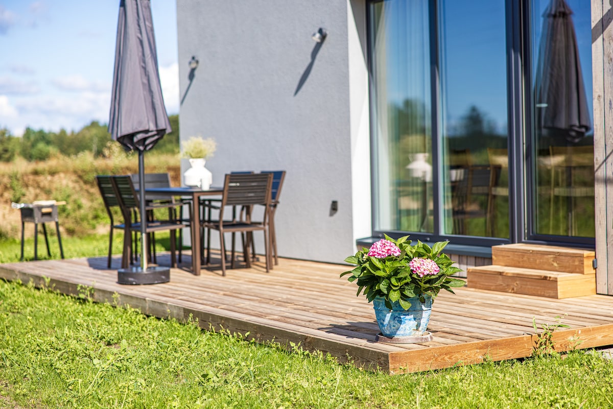 A wood deck area is presented, featuring a dining set with four chairs and a table. A large umbrella provides shade. A potted plant with vibrant flowers is positioned near the entrance, and large glass doors offer a view of the surrounding green landscape.