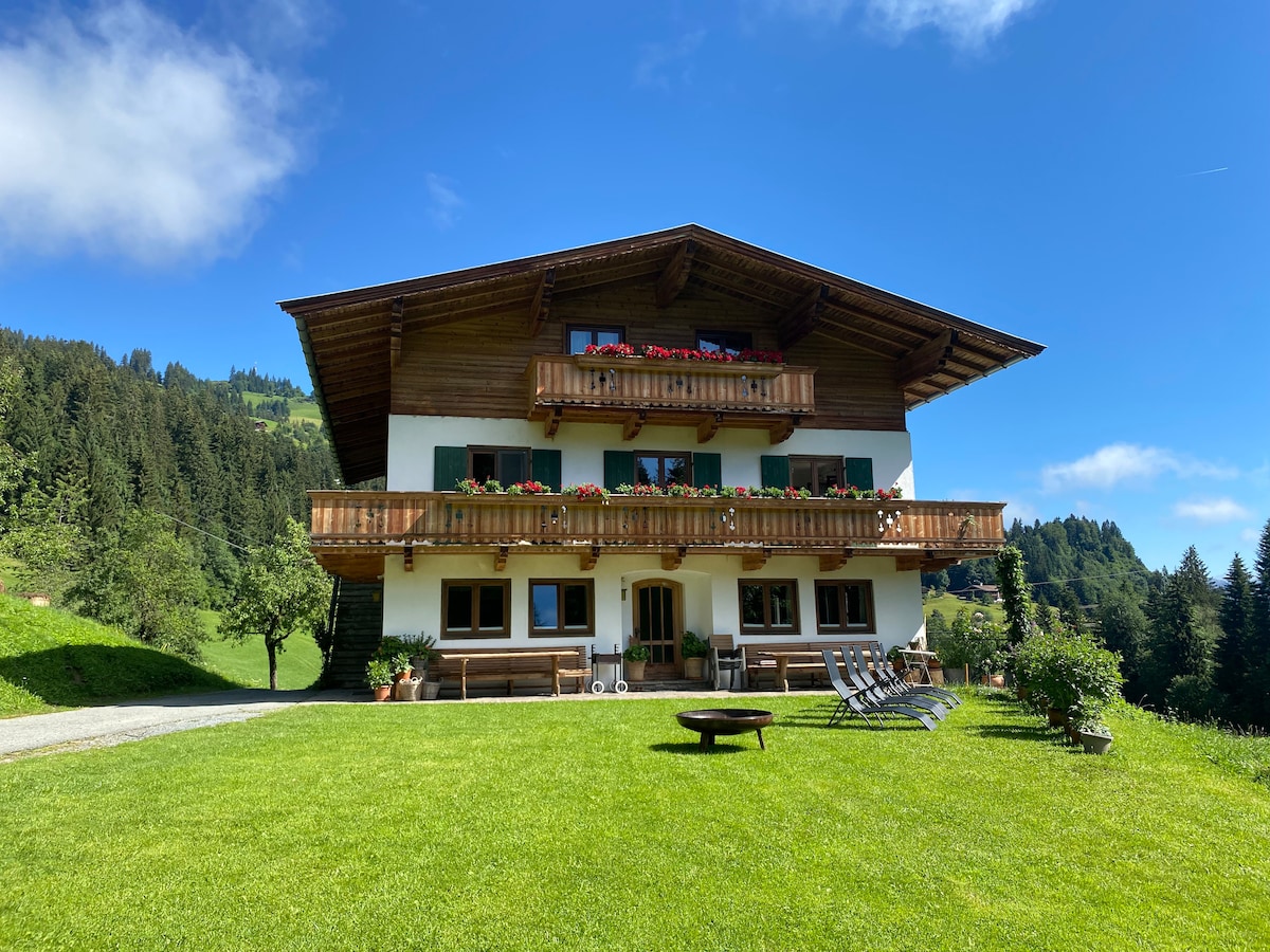 A charming mountain farmhouse is set against a clear blue sky, featuring multiple balconies adorned with flower boxes. A well-manicured lawn extends before the building, with benches and lounge chairs available for guest use. Surrounding greenery and trees enhance the serene environment.