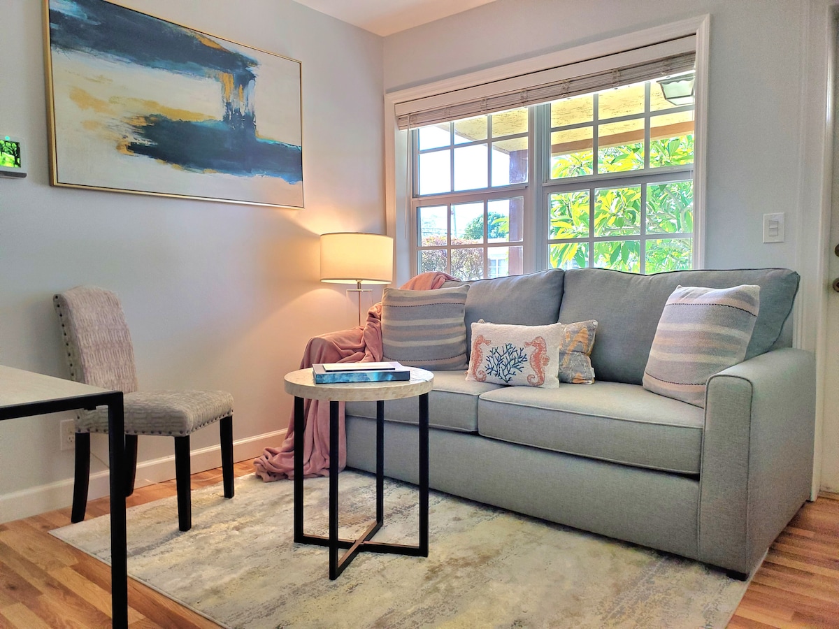 A comfortable seating area is highlighted by a cozy gray sofa accented with decorative pillows. A small round table sits in front, accompanied by a stylish chair. Natural light streams in through large windows adorned with soft curtains, offering views of greenery outside.
