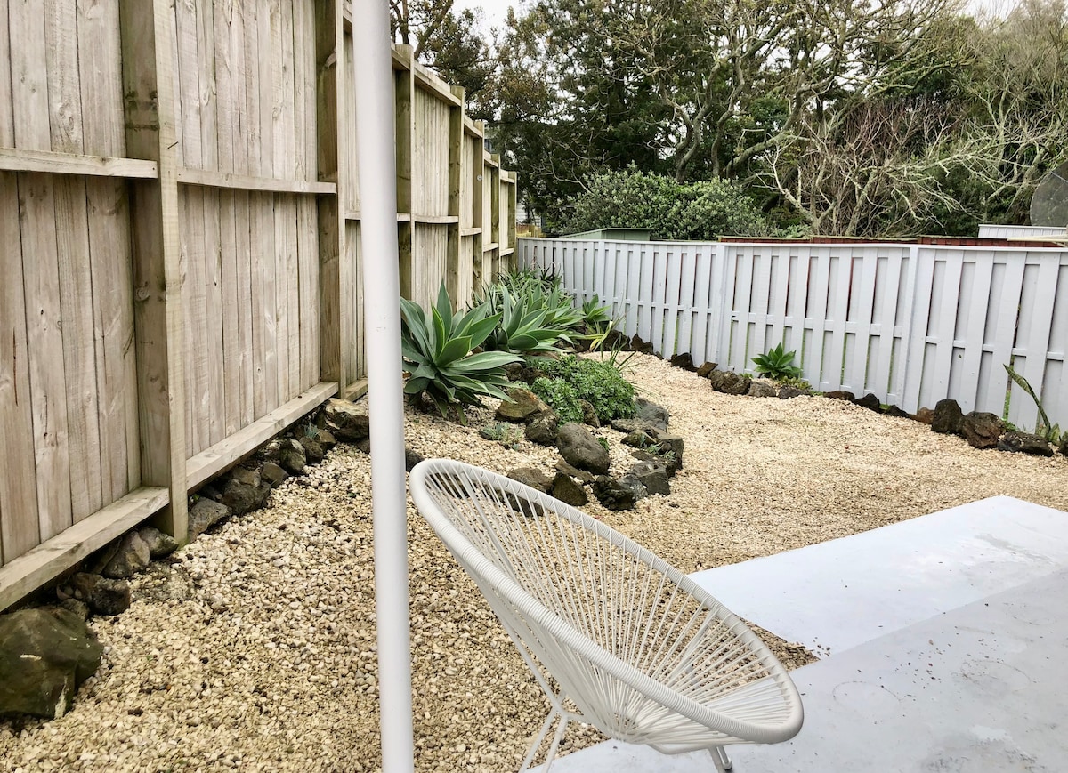 A scenic fenced courtyard is revealed, featuring a minimalist landscape design with gravel and strategically placed plants. A comfortable white chair sits on a patio, offering a quiet space to relax outdoors. Lush greenery can be seen along the perimeter, adding natural beauty.