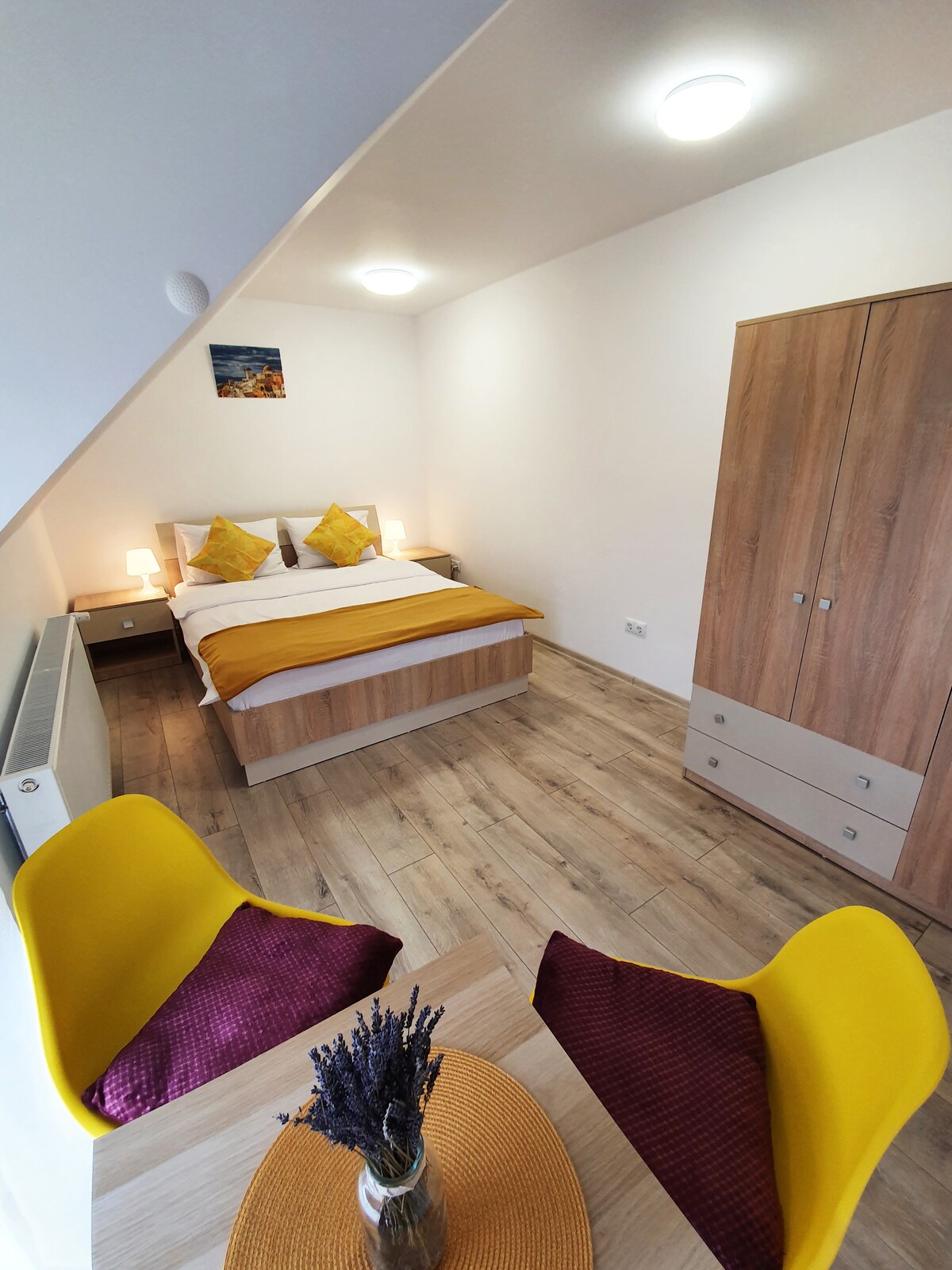 A cozy bedroom features a spacious bed with a warm yellow accent blanket. Warm lighting is provided by bedside lamps on either side. A standard closet stands against the wall, while a small table with two yellow chairs adds a touch of comfort to the space.