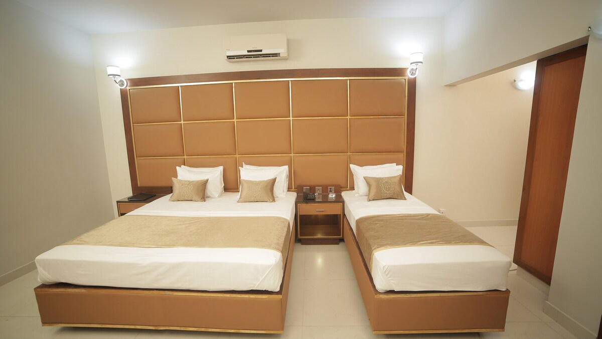A well-appointed room features two spacious beds with neutral tones and elegant headboards. Soft bedding is complemented by accent pillows, and warm lighting enhances the modern decor. A nightstand with lamps is positioned between the beds, contributing to the room's functional layout.