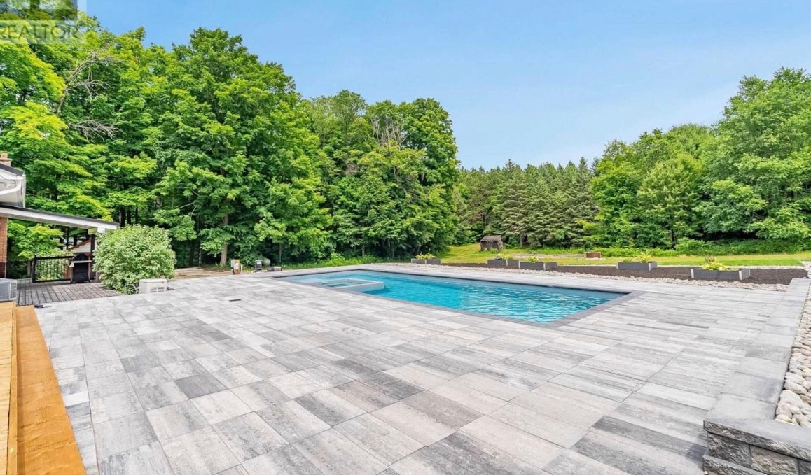 An in-ground swimming pool is surrounded by a stone patio, nestled within a lush green landscape. Trees border the area, providing a serene backdrop. Sunlight reflects off the clear water, inviting relaxation in a tranquil outdoor setting.
