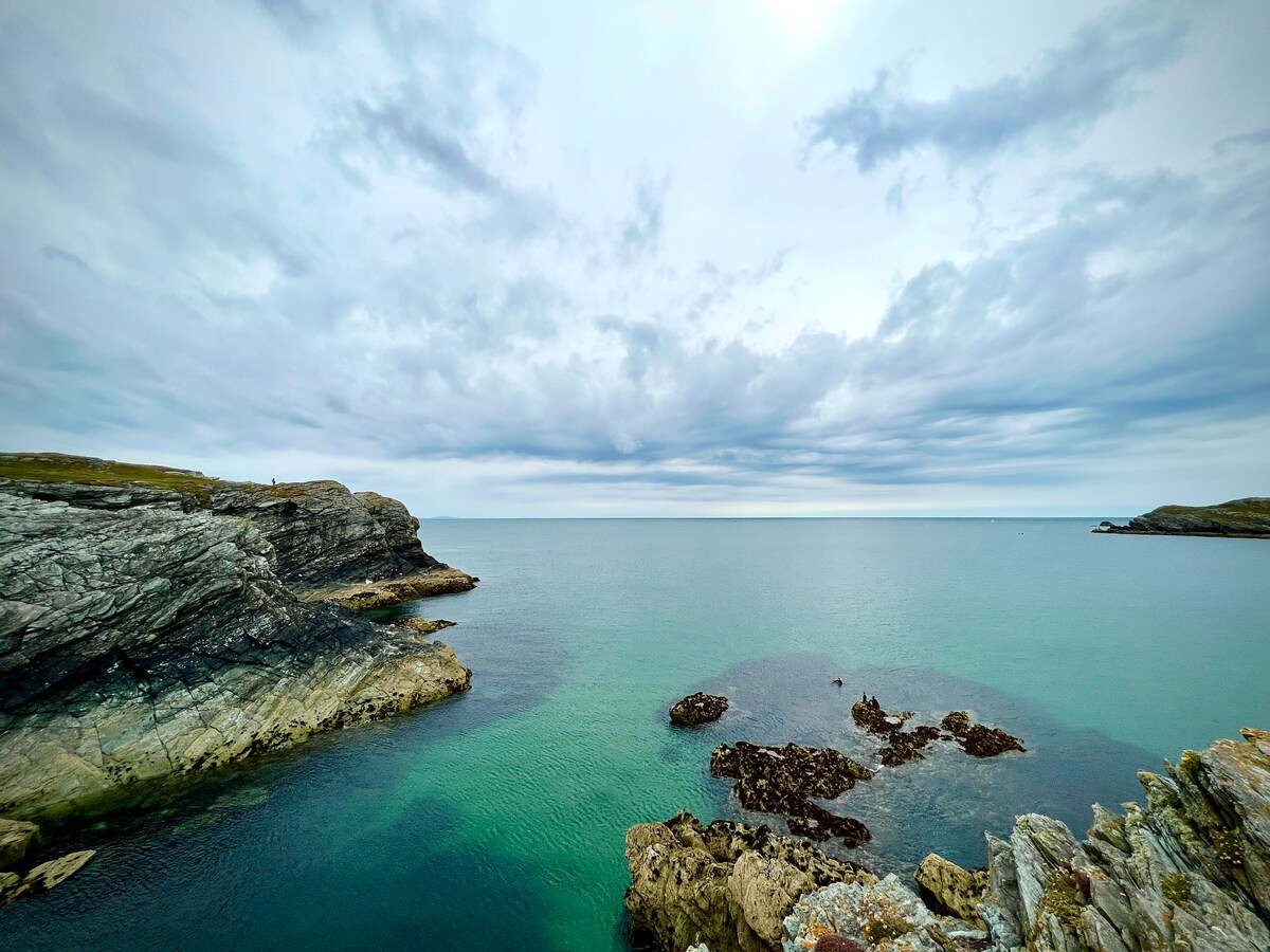 The image captures a serene coastal scene with calm waters reflecting shades of turquoise and gray. Rocky cliffs frame the shoreline, while distant land is visible under a cloudy sky. A small group of rocks emerges from the water, inviting exploration.