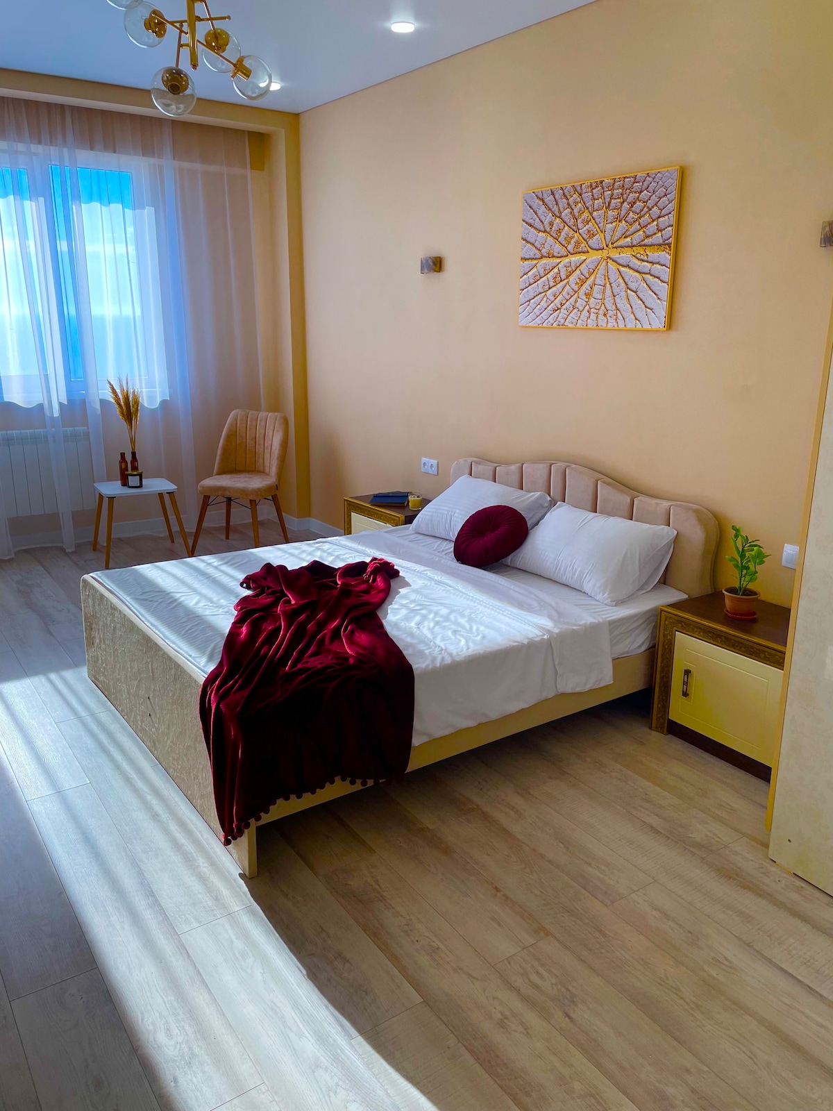 A bright sleeping area features a spacious bed with white linens and a burgundy throw blanket casually draped over the side. A modern chair and a small table are positioned near a window, which provides natural light. Decorative wall art adds a touch of style.