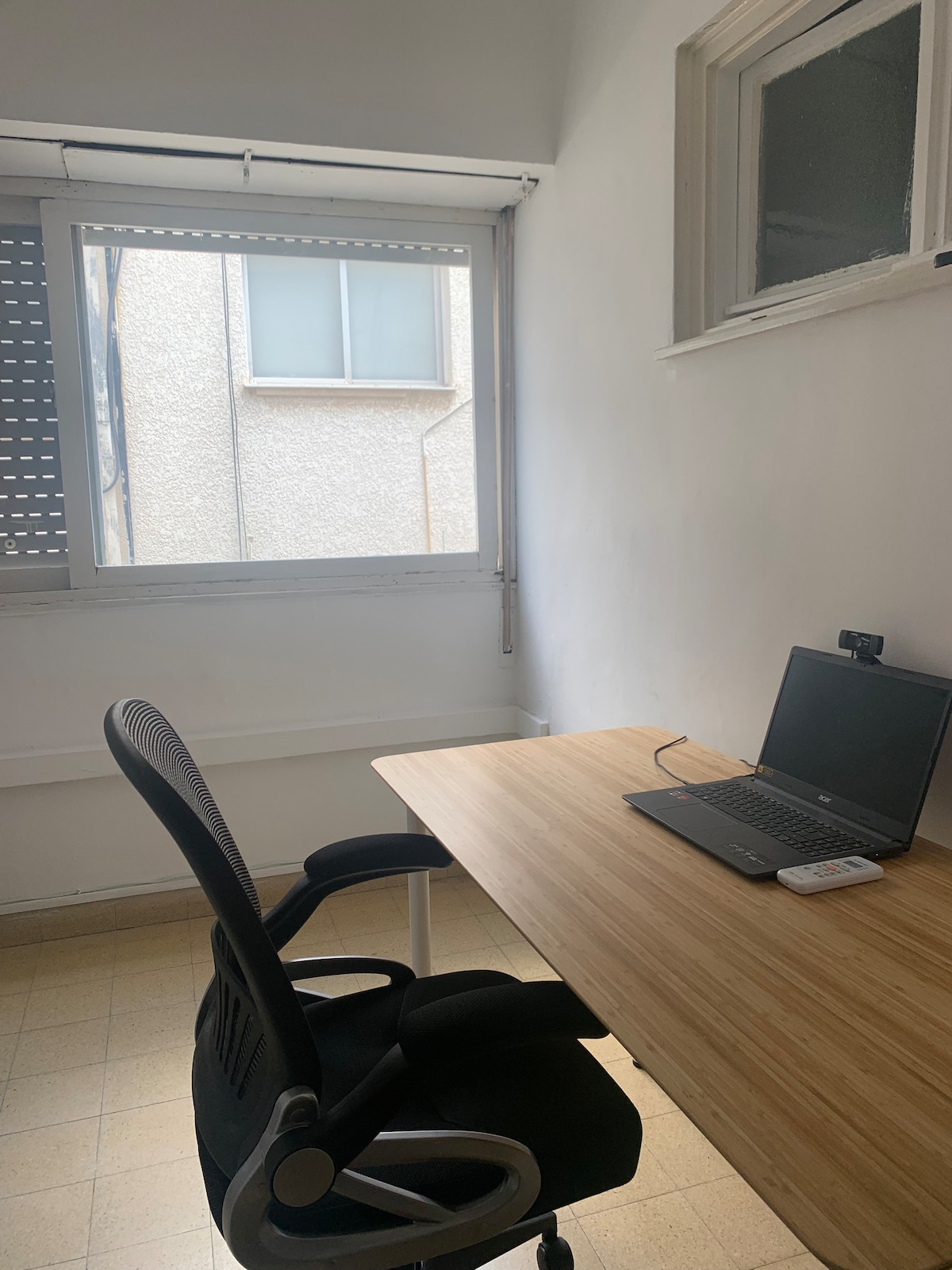 A workspace area features a wooden desk with a laptop, accompanied by an ergonomic black chair. Natural light enters through a window, which has privacy blinds partially lowered. A neutral wall complements the simple and functional arrangement.