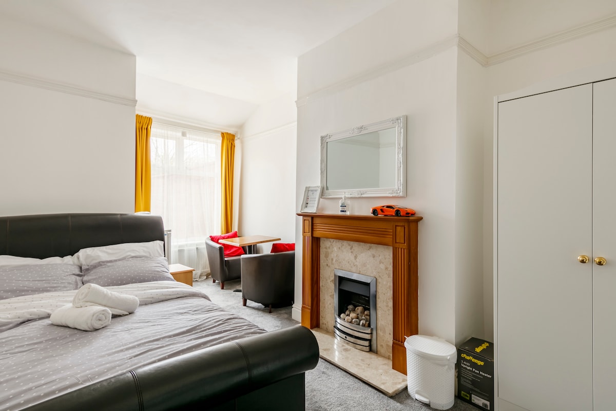 A private room features a spacious bed covered with a light blanket, paired with a fireplace and a large mirror above a wooden mantel. Two armchairs are situated nearby, with bright curtains framing a window that allows natural light to fill the room.