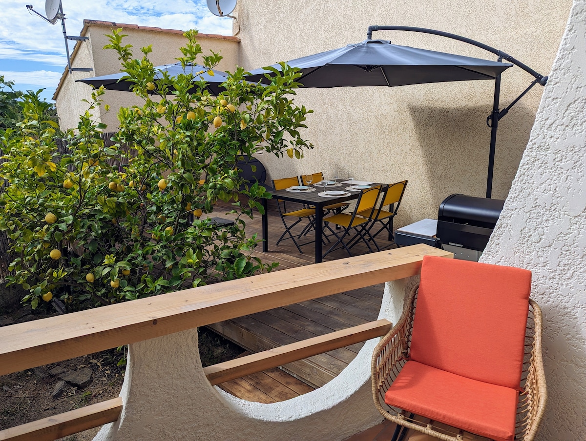A spacious terrace is shown, featuring a dining set with yellow chairs and a black barbecue. A lemon tree provides green foliage and fruit. An orange chair is positioned nearby, framing the view of the outdoor space beneath a shaded area created by large umbrellas.
