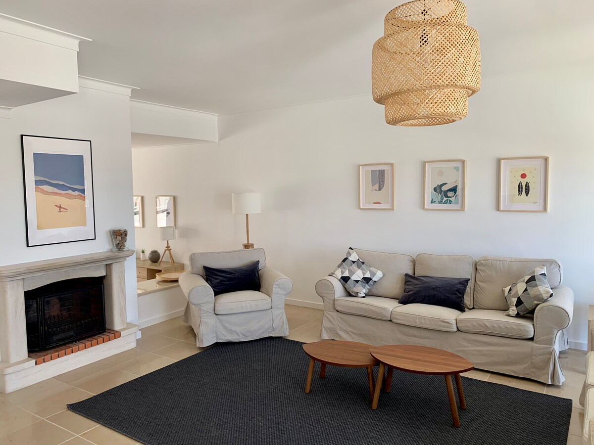The spacious living room features a comfortable light-colored sofa and armchair, complemented by decorative cushions. A woven light fixture hangs above, while a pair of wooden coffee tables sit on a textured area rug. Framed artwork adorns the wall, enhancing the tranquil atmosphere.