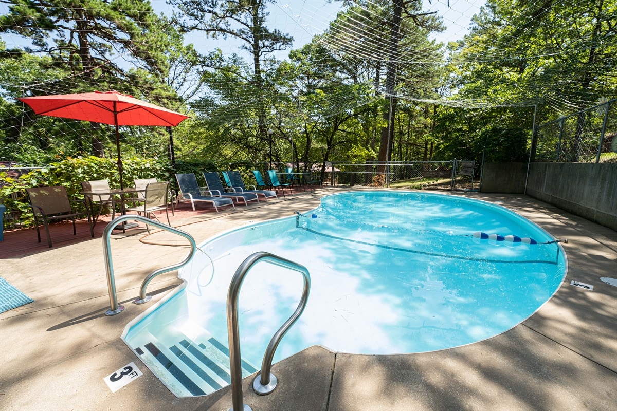 A clear blue pool is surrounded by tall trees, providing a serene environment. Nearby, a shaded area features several lounge chairs and a vibrant red umbrella. Steps lead into the pool, which is marked for depth near the edge.