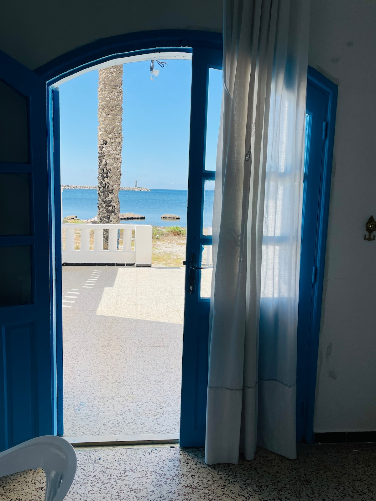 A bright blue door opens to a scenic view of the sea, with a palm tree visible in the foreground. Sunlight filters through sheer curtains, illuminating the entryway and creating a welcoming atmosphere.