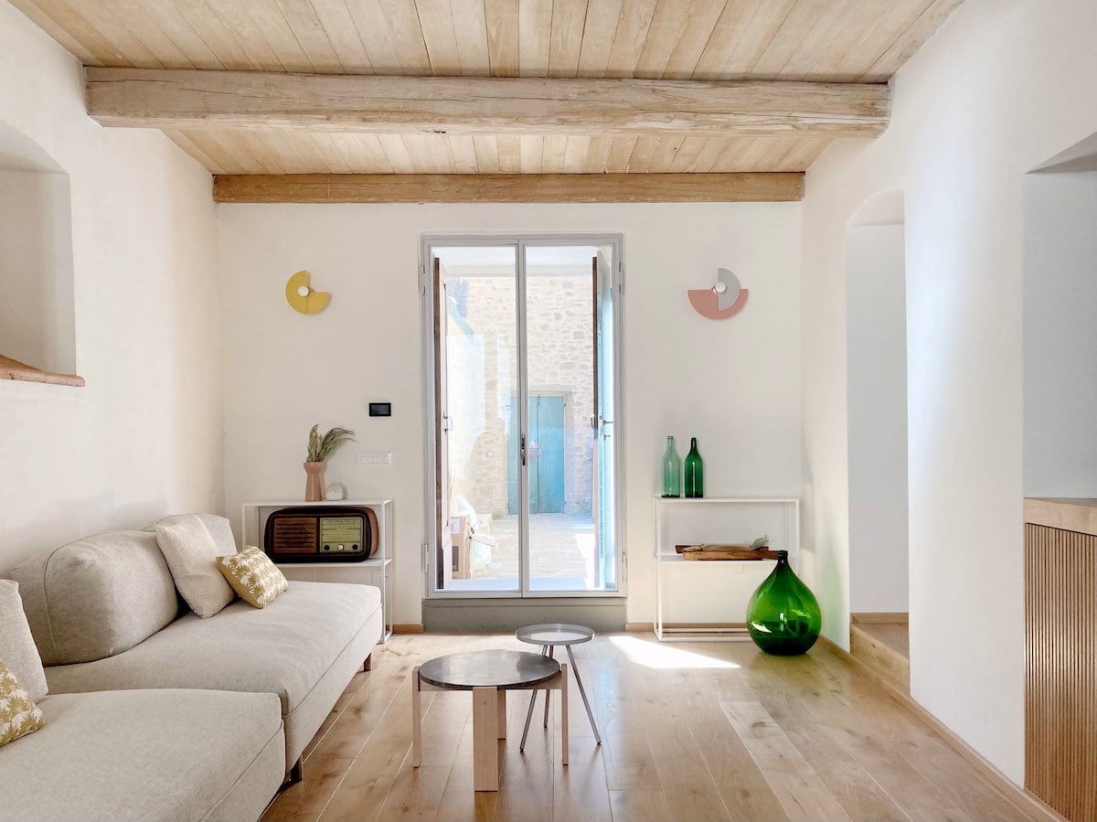 A light-filled living room features a cozy, L-shaped couch adorned with decorative cushions. A small round table sits between two green glass vases, while glass doors lead to an outdoor area. The wooden ceiling and neutral tones create a serene ambiance.