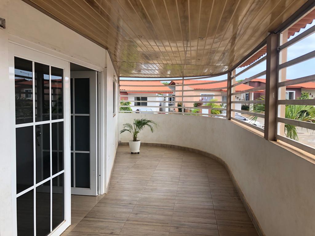 A covered terrace features smooth wooden flooring and large windows that allow natural light to brighten the space. Potted plants are positioned around the area, creating a serene outdoor retreat adjacent to the interior living spaces.
