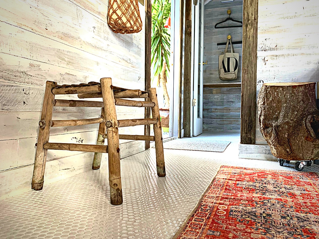 A rustic entrance area is highlighted by a handcrafted wooden stool and a vintage-inspired area rug. A door opens toward a pathway lined with plants, while a bag hangs on a hook, adding to the cozy ambiance of the space.