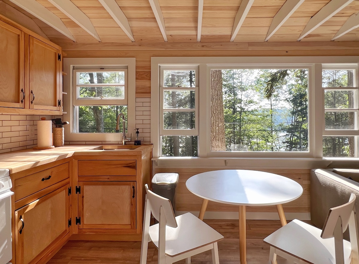 The kitchen area features wooden cabinetry with ample counter space. A round white table is surrounded by two chairs, positioned beneath large windows that provide a view of the surrounding trees and the water. Natural light floods the space, enhancing its warm atmosphere.