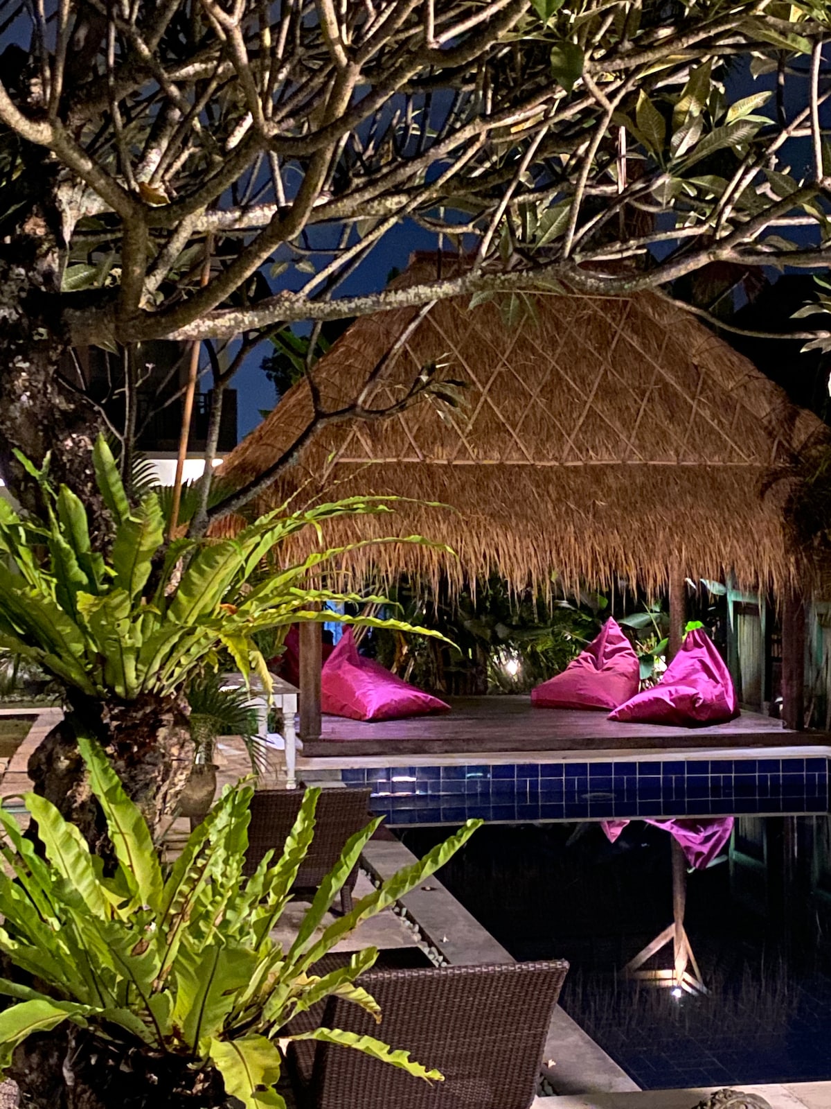 A thatched gazebo is visible beside a pool, featuring colorful bean bags for seating. Lush greenery surrounds the area, enhancing the tropical feel. Soft lighting highlights the gazebo and is reflected in the calm water of the pool, creating a relaxing atmosphere.