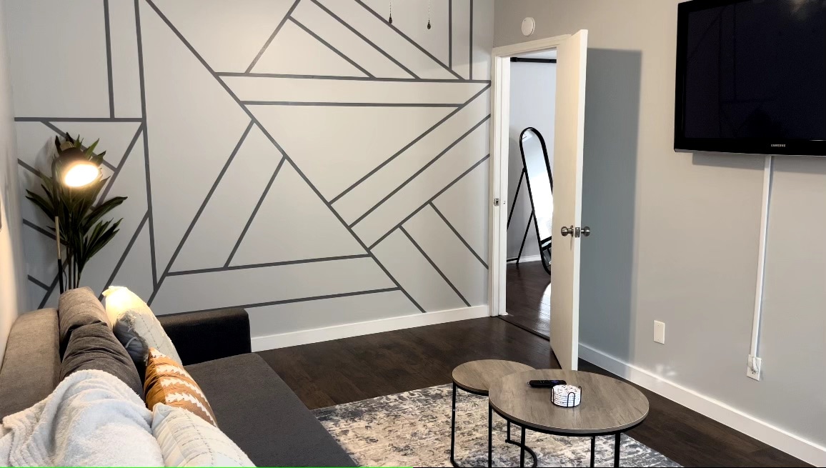 A modern living space features a minimalistic design with geometric wall accents. A comfortable sofa is positioned in front of a coffee table, while a wall-mounted TV offers entertainment options. A doorway leads to additional private areas, enhancing the spacious feel.