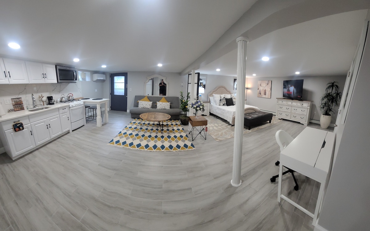 The spacious, newly renovated living area features a combination of a cozy sleeping space with a large bed and a dining area. A modern kitchen is visible, alongside a comfortable work desk and ample natural light from nearby windows. Decorative plants and rugs add warmth.