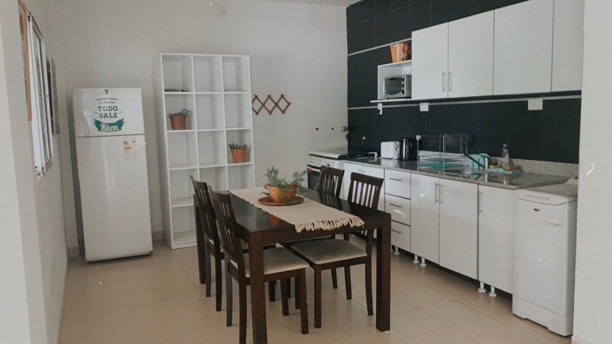 A modern kitchen and dining area is featured, showcasing a sleek design with white cabinetry and a dark accent wall. A wooden dining table is surrounded by six chairs, and a refrigerator is positioned nearby. Natural light spills into the space, illuminating the neutral tile flooring.